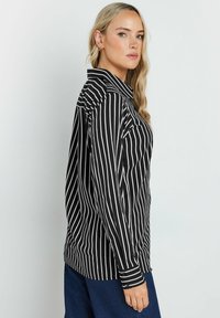 Camicia a righe bianco e nere con colletto, dotata di maniche lunghe e una vestibilità leggermente ampia, realizzata in tessuto leggero.