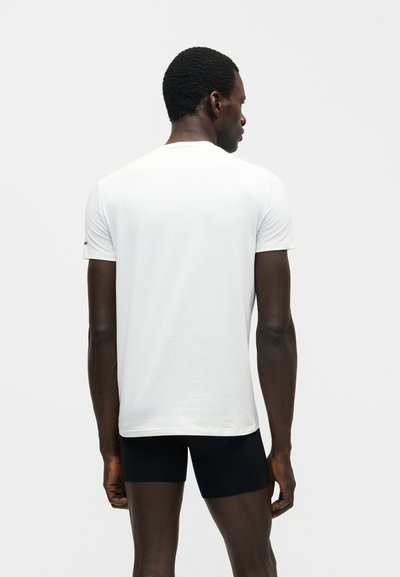 Άνδρας που είναι γυρισμένος πλάτη, φοράει ένα απλό λευκό κοντομάνικο μπλουζάκι και μαύρα στενά σορτσάκια σε λευκό φόντο.