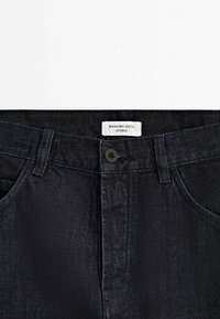 Tumma denimi-farkut sileällä pinnalla, joissa on edessä nappikiinnitys ja etiketti, jossa lukee "Massimo Dutti Studio." Klassinen muotoilu.