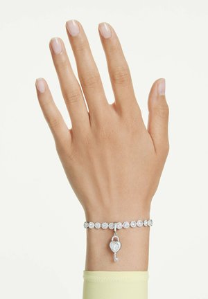 Roka, ki nosi srebrno teniško zapestnico s krožnimi kamni in obeskom v obliki ključa v obliki srca, na svetlem enobarvnem ozadju.