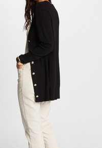 Cardigan noir à manches longues avec une coupe décontractée, présentant des boutons décoratifs sur le côté, associé à un pantalon beige clair.