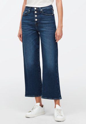 Kvinde iført højtaljede, mørkeblå bukser med vide ben og knaplukning samt hvide sneakers på hvid baggrund.