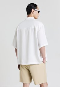 Uomo che indossa occhiali da sole neri, una camicia bianca a maniche corte con bottoni, pantaloni corti beige, in piedi con le mani nelle tasche, voltato di spalle.