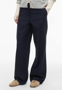 Pantalon large bleu marine en tissu respirant, doté d'une fermeture éclair à l'avant et de poches latérales, avec une texture lisse et un design ajusté.