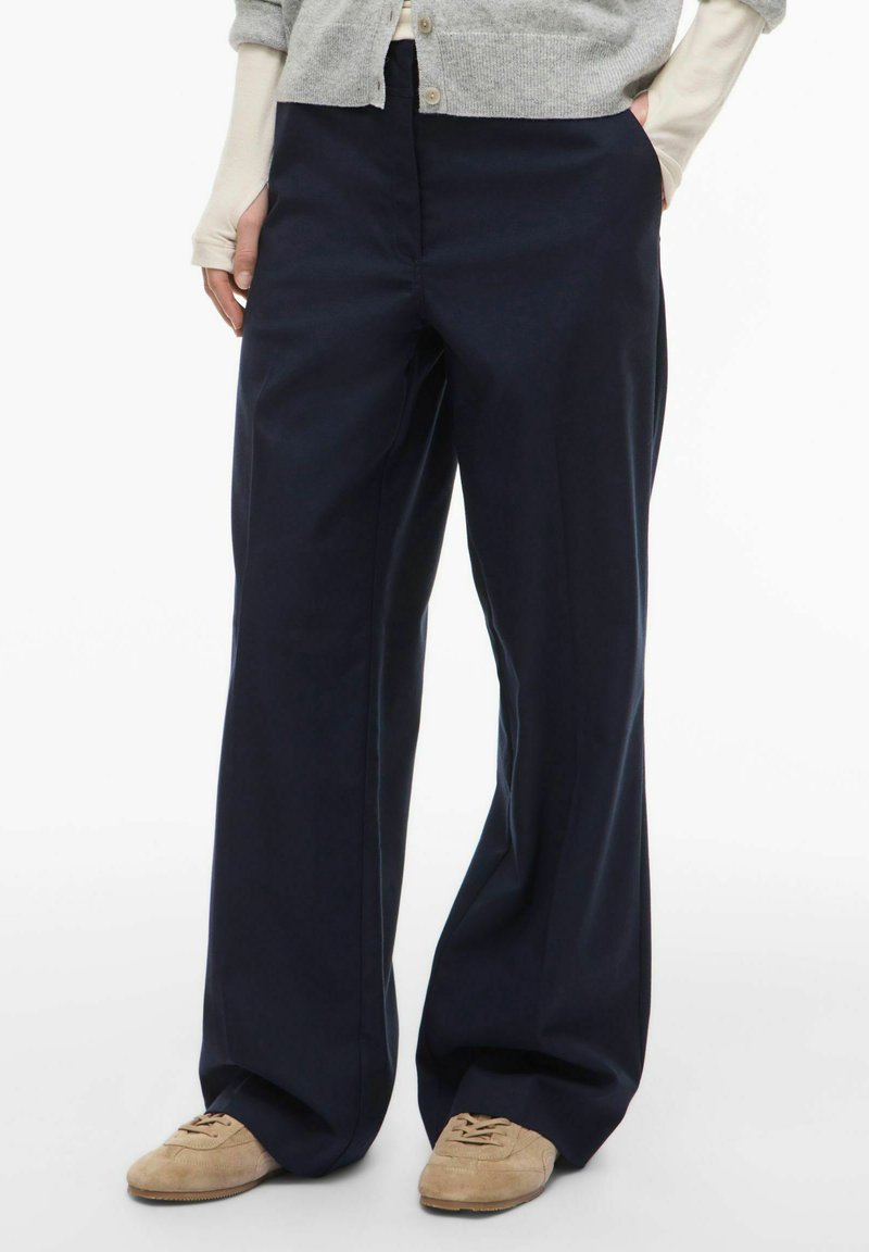 Pantalon large bleu marine en tissu respirant, doté d'une fermeture éclair à l'avant et de poches latérales, avec une texture lisse et un design ajusté.