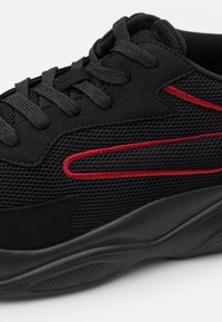 Baskets noires avec une tige en mesh et des accents en daim. Détail rouge le long du côté, semelle en caoutchouc noire robuste avec une adhérence texturée.