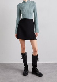 Světlá tyrkysová žebrovaná košile s dlouhým rukávem, černá áčková sukně a černé vysoké boty na šněrování. Hladký povrch, přiléhavý střih a strukturovaná silueta.