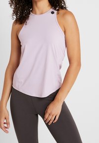 Světle fialová sportovní tílko s vysokým výstřihem a malým černým logem na rameni. Disponuje měkkou a hladkou látkou a uvolněným střihem.