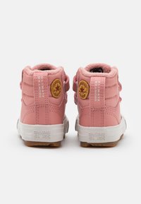 Zapatillas altas rosas con parte superior de tela texturizada, suelas de goma y un parche con logo circular. Incluyen correas de velcro para facilitar su uso.