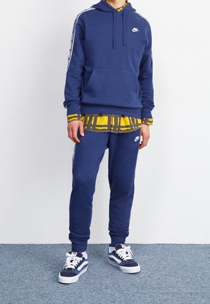Personne portant un survêtement Nike bleu marine avec des logos blancs, un t-shirt jaune et noir à motifs en dessous, et des baskets noires et blanches.