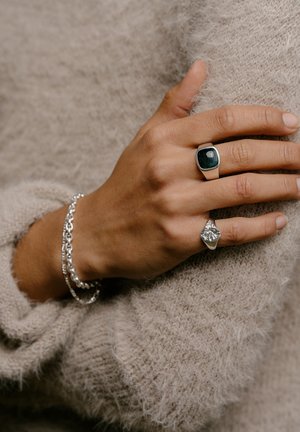 Sølv ringe med mørkegrønne og klare stenaccenter vist på en hånd, prydet med et sølv kædearmbånd mod en struktureret beige sweater.