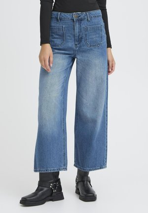 Weite blaue Denim-Jeans mit hoher Taille, ausgestattet mit zwei Fronttaschen und zwei Gesäßtaschen, kombiniert mit schwarzen Knöchelstiefeln.