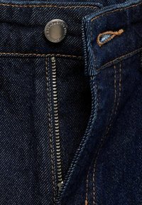 Gros plan sur un jean en denim bleu foncé avec une fermeture éclair argentée partiellement ouverte et un bouton métallique au-dessus avec des détails de couture orange.