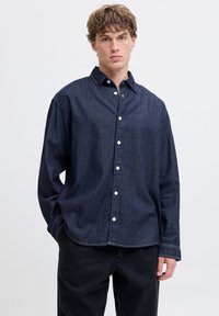 Mørkeblå denimskjorte med knappestolpe foran, spredt krage, lange ermer og en avslappet passform. Har hvite knapper og kontrasterende søm.