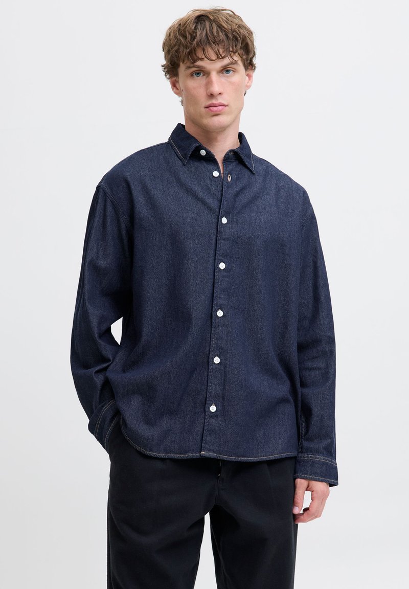 Mørkeblå denimskjorte med knappestolpe foran, spredt krage, lange ermer og en avslappet passform. Har hvite knapper og kontrasterende søm.