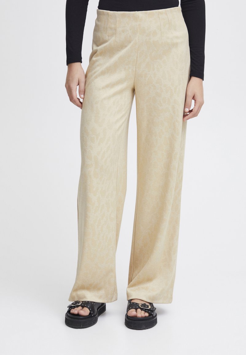 Wijduitlopende broek van zacht beige stof met een subtiel dierenprintpatroon. Heeft een hoge taille en een soepele textuur. Gecombineerd met zwarte sandalen.