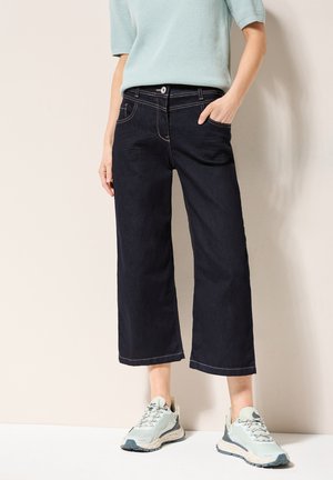 Culottes de mezclilla oscura con un corte de pierna ancha, que presentan costuras de contraste, bolsillos y una cintura alta, combinados con zapatillas de color claro.