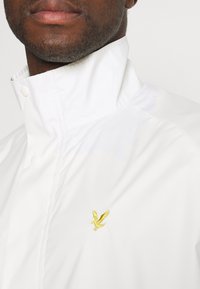 Zbliżenie na osobę w białej kurtce z podniesionym kołnierzem i małym żółtym logo orła na piersi.