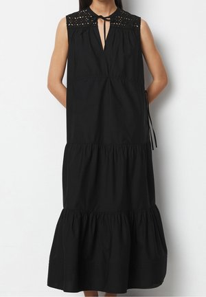 Robe midi noire sans manches avec jupe à volants, empiècement en dentelle ajourée et col à nouer portée par une personne debout les mains dans le dos.