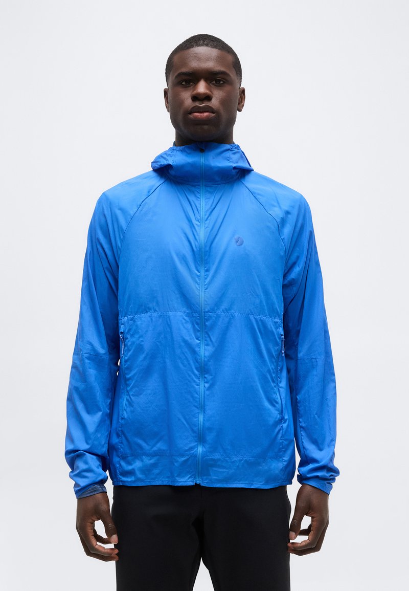 En ung mand iført en knaldblå lynlås-windbreaker med hætte, stående mod en ensfarvet lys baggrund og vendt mod fronten.