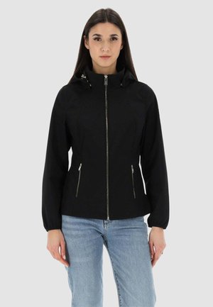 Donna che indossa una giacca nera con cerniera, cappuccio e tasche con cerniera, abbinata a jeans azzurri, in piedi davanti a uno sfondo semplice.