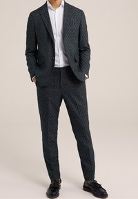 Muž v tmavém vzorovaném obleku s bílou košilí a černými brusinkovými mokasínami, stojící s rukama v kapsách na jednobarevném pozadí.