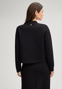 Sweatshirt noir à manches longues, coupe décontractée et col rond. Présente un accent logo argenté au dos du col. Texture lisse.