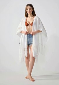 Bílý kimono střihu s třásněmi, vyrobený z lehké látky s otevřenými rukávy, nošený přes hnědé bikiny a džíny šortky.