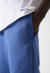 Pantaloni de trening albaștri, cu o textură netedă, prezintă un logo pe lateral. O cămașă albă cu un etichetă logo albastră este vizibilă deasupra pantalonilor.