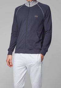 Man wearing navy zip-up jacket with gray collar and light gray textured pants, both branded with BOSS Hugo Boss logos.