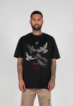 Sort bomulds T-shirt med grafik af duer og kæder, med ordet "FREEDOM" i rødt. Korte ærmer og en afslappet pasform.