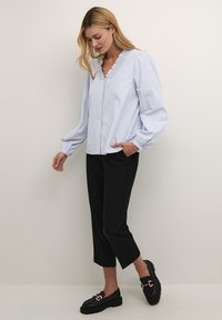 Weißes Hemd mit blauen Streifen, Rüschensaum und Knöpfen; kombiniert mit schwarzen, cropped Hosen und schwarzen Loafers mit goldenen Beschlägen.