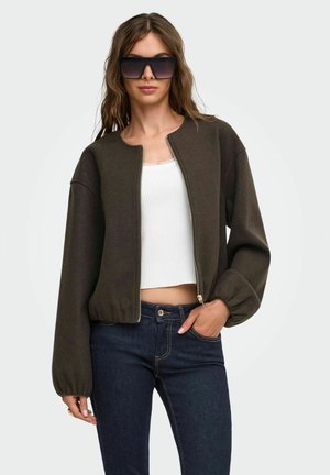 Femme portant de grandes lunettes de soleil noires, une veste marron foncé zippée, un haut court blanc et un jean bleu foncé, posant avec une main dans la poche.