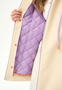 Ręka trzyma otwarty beżowy płaszcz, ukazując pikowaną jasnofioletową podszewkę, pomarańczowe zatrzaski oraz fioletowe wykończenia z sznurkiem na rękawie.
