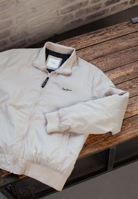 Beige bomberjacka med dragkedjestängning, ribbade muddar och krage. Har en subtil logotyp på bröstet och en mjuk, glänsande tygstruktur.