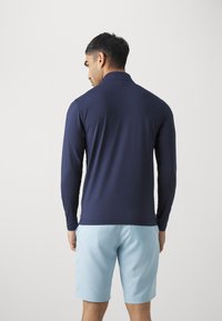 Marineblå langermet skjorte med krage, båret med lyseblå shorts. Glatt tekstur, minimalistisk design, enkel passform. Bakview.