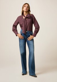 Bordeaux geruite blouse met een strikdetail, gecombineerd met hoogriem blauwe jeans met studversieringen aan de zijkanten.