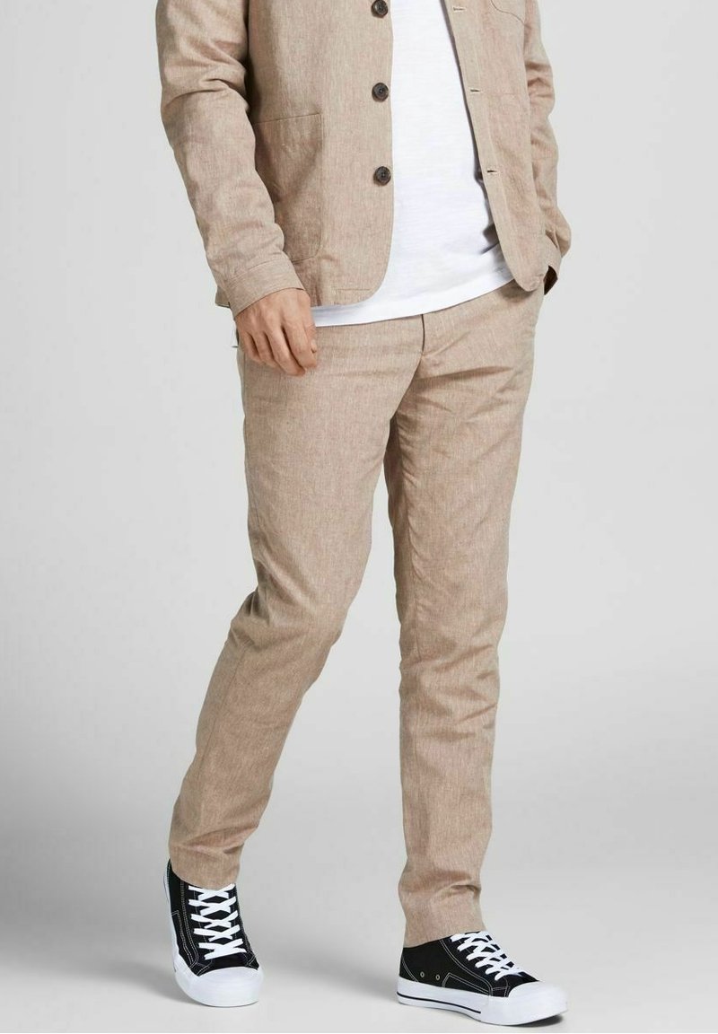 Veste de costume en lin beige avec deux poches et des boutons foncés, assortie à un pantalon de costume et des baskets noires avec des lacets blancs.
