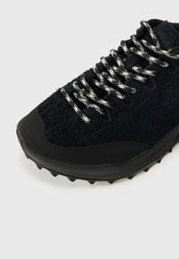 Čierna teniska s textúrovaným zvrškom, vybavená čiernymi gumovými detailmi a čierno-bielymi šnúrkami s vzorom. Podrážka so vzorkou na lepšiu priľnavosť.