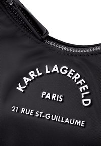 Fekete szövet táska fényes cipzárral, kidomborodó fehér szöveggel: "KARL LAGERFELD," "PARIS," és "21 RUE ST-GUILLAUME" íves elrendezésben.