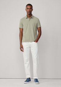 Polo shirt verde chiaro, maniche corte, colletto, abbinato a jeans bianchi e scarpe da ginnastica blu, in piedi contro uno sfondo neutro.
