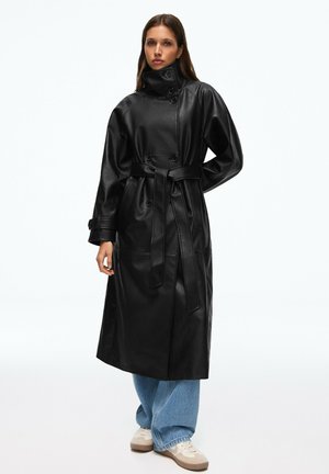 Frau trägt einen schwarzen, taillierten Ledertrenchcoat, blaue, weit geschnittene Jeans und weiße Sneaker, steht mit einer Hand hinter dem Rücken.