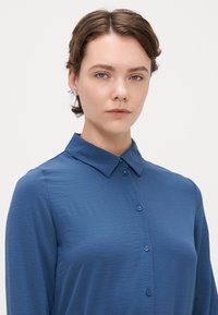 Μπλε πουκάμισο με κουμπιά, φτιαγμένο από υφασμάτινη, ελαφριά υφή, με κοφτό γιακά και μακριά μανίκια, διαθέτοντας ταιριαστά κουμπιά κατά μήκος του μπροστινού μέρους.