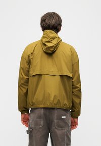 Άνδρας που φοράει αθλητικό μπουφάν με κουκούλα σε ελιά πράσινο χρώμα και γκρι παντελόνι εργασίας, φωτογραφημένος από πίσω σε απλό λευκό φόντο.