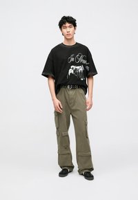 Melna pārliela izmēra T-krekls ar grafisko apdruku, kombinēts ar olīvu zaļas krāsas kabatām bagātām cargo biksēm. Melni apavi pabeidz tēlu.