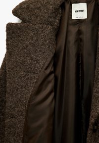 Abrigo marrón con textura y exterior suave. El interior cuenta con un forro liso de color marrón oscuro. La etiqueta muestra el nombre de la marca "Koton."