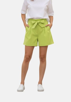 Donna che indossa pantaloncini verde lime a vita alta con cintura annodata e scarpe bianche casual, in piedi con le mani nelle tasche.