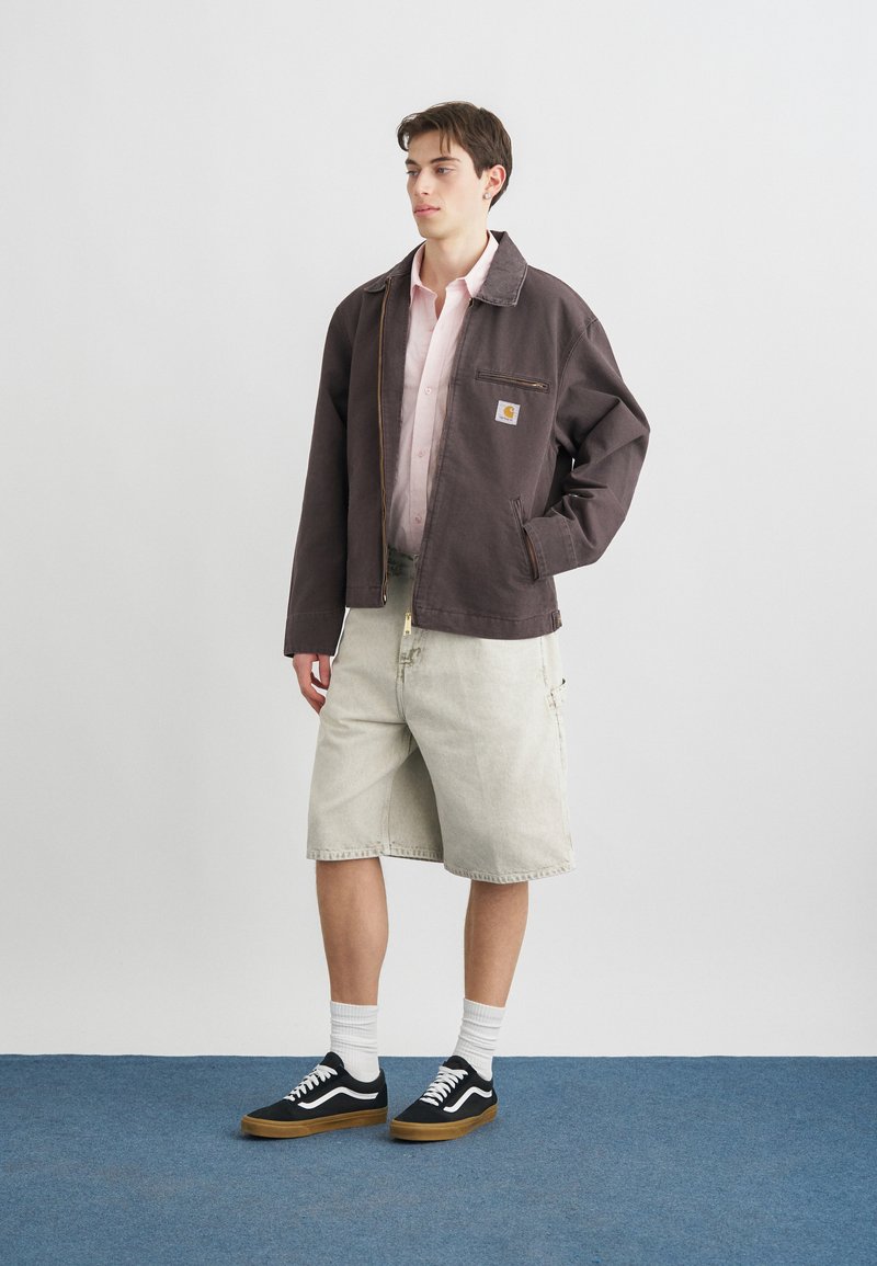 Joven de pie con chaqueta de trabajo marrón oscuro, camisa rosa claro, pantalones cortos beige hasta la rodilla, calcetines blancos y zapatillas negras sobre suelo azul.