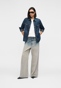 Dunkelblaues Jeanshemd mit zwei aufgesetzten Brusttaschen und Knopfleiste, kombiniert mit grauer, weit geschnittener Hose aus strukturiertem Stoff.