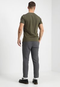 Muž stojící zády, oblečený v olivově zeleném tričku, šedých kalhotách, bílých ponožkách a černých botách v prostorné bílé místnosti.